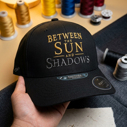 A close-up view of the black baseball cap, with the text 'BETWEEN THE SUN AND SHADOWS' embroidered on the front.
